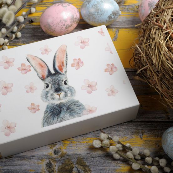 Eine weiße Box mit einer Hasenillustration ist von Ostereiern und floralen Elementen auf einer Holzoberfläche umgeben. Der Hase ist grau mit rosa Blumen um ihn herum.