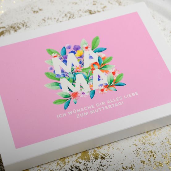 Eine weiße Schachtel mit einem rosa Deckel zeigt ein florales Design mit 'MAMA' in der Mitte und 'Ich wunsche dir alles Liebe zum Muttertag!' darunter. Das Design ist vor einem rosa Hintergrund angebracht.