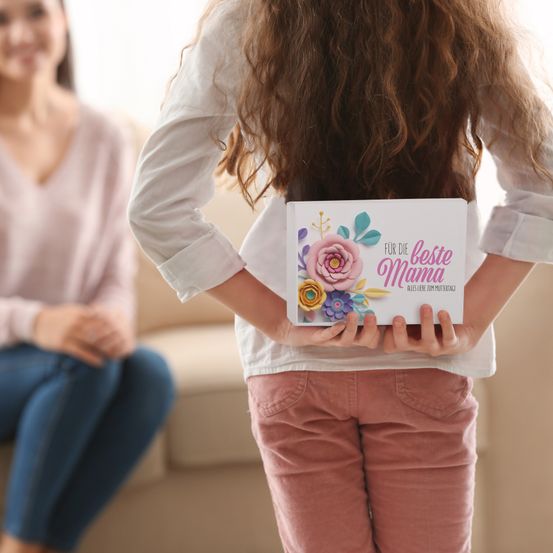 Ein Mädchen hält eine Schachtel mit der Aufschrift 'Für die beste Mama' und steht mit dem Rücken gedreht. Im Hintergrund sitzt eine andere Frau auf einem Sofa.