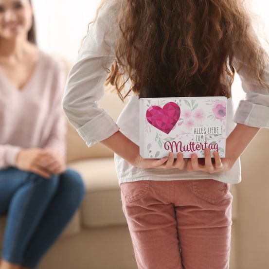 Ein Mädchen hält eine Karte mit Herz-Design und Blumenmustern und zeigt 'Alles Liebe zum Muttertag'. Eine Frau sitzt auf einer Couch im Hintergrund.