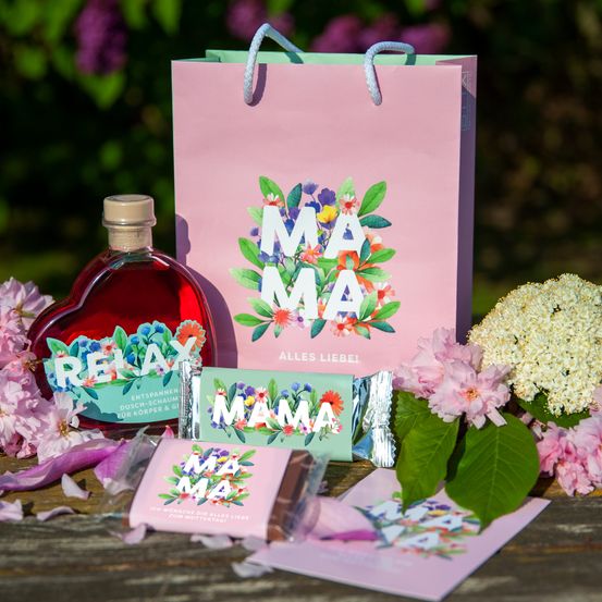 Ein rosafarbener Papierbeutel mit dem Aufdruck 'Mama', eine Flasche roten Sirups mit 'Entspann dich' und kleine Schokoladen mit 'Mama' liegen auf einem Tisch mit Blumen.