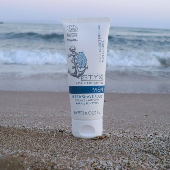 Eine Tube Männer-Rasierwasser auf dem Strand mit Wellen im Hintergrund. Das Produkt ist von Styx und ist vegan.