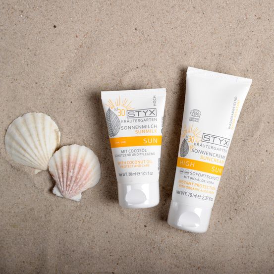 Zwei Tuben Sonnencreme mit Kokosöl liegen neben zwei Muschelschalen auf dem Sand. Eine Tube ist mit 'SUN' und die andere mit 'HIGH' beschriftet.