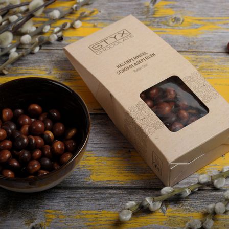 Ein mit schokoladeüberzogenen Haselnüssen gefüllter Becher steht neben einer braunen Pappschachtel mit der Aufschrift 'Hasenpemmerl Schokoladeperlen' auf einer gelben Holzoberfläche.