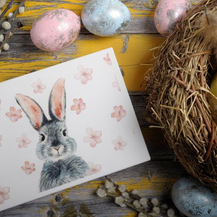 Auf einem Holztisch befindet sich eine Karte mit einer Hasenillustration, umgeben von Ostereiern, Blumen und einem Nest. Die Eier haben Pastellfarben und Blumenmuster.