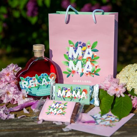 Eine herzförmige Flasche mit rotem Sirup, ein rosa Geschenktüte mit 'MAMA', rosa Blumen und einem Geschenkanhänger. Ein blumengeschmücktes Papier und eine Karte mit 'MAMA'. Ein rosa Geschenkanhänger mit 'ALLES LIEBEL'.