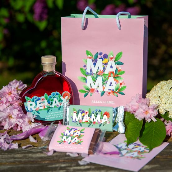 Eine herzförmige Flasche mit rotem Sirup, ein rosa Geschenktüte mit 'MAMA', rosa Blumen und einem Geschenkanhänger. Ein blumengeschmücktes Papier und eine Karte mit 'MAMA'. Ein rosa Geschenkanhänger mit 'ALLES LIEBEL'.
