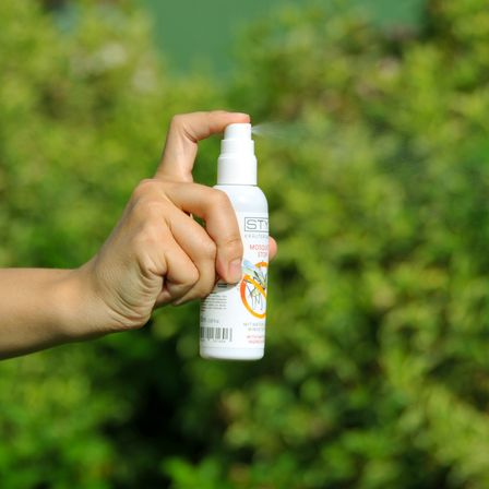 Eine Hand hält eine weiße Sprühflasche mit einem Aufkleber, der 'Moskito-Spray' und ein Bild eines Moskitos zeigt. Der Spray wird im Freien verwendet.