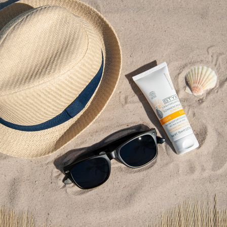 Ein Hut, Sonnenbrille, Sonnencreme und Muschelschale auf sandigem Strand. Der Hut hat einen blauen Streifen.