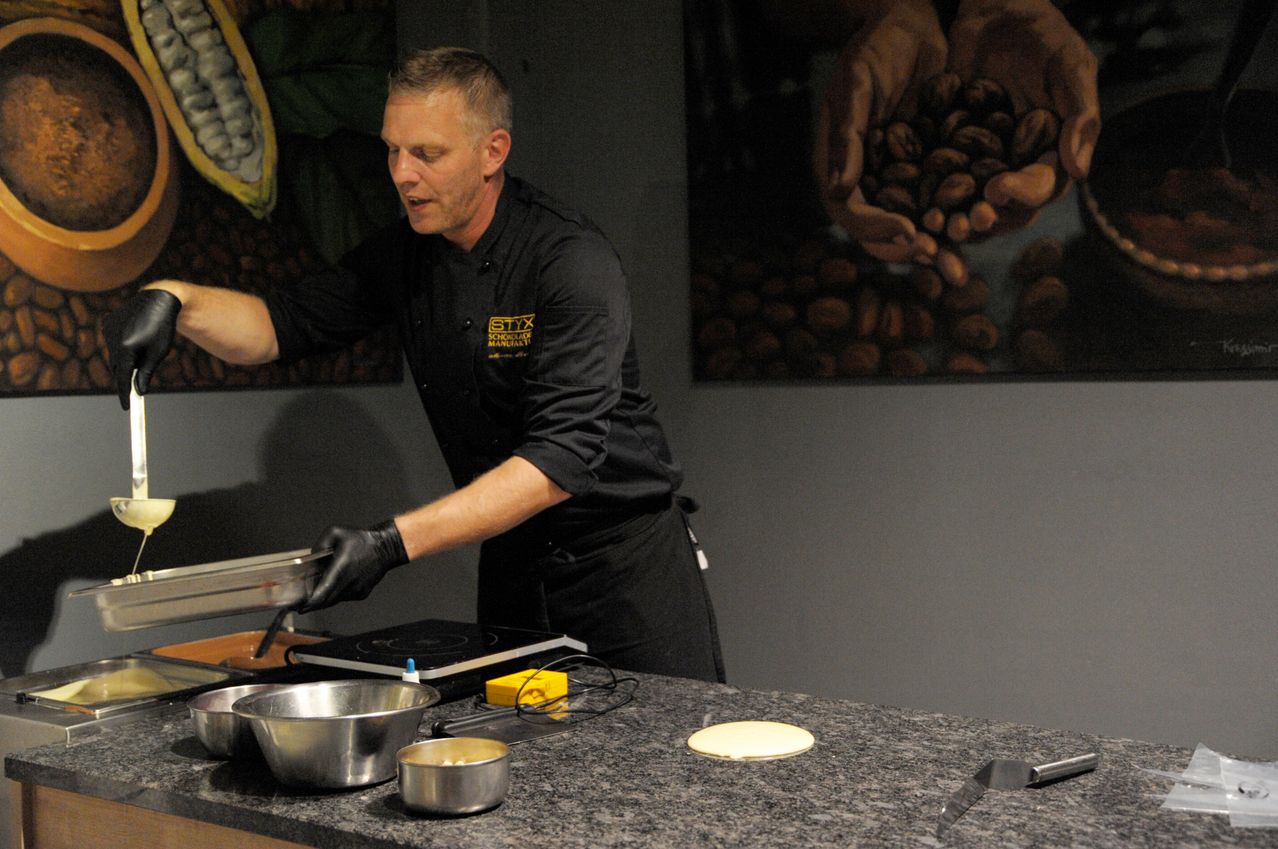 Ein Koch kocht in einer Küche mit Granitarbeitsplatte. Er trägt ein schwarzes Kochuniform und Handschuhe. Es gibt Schüsseln, eine Pfanne und ein Messer auf der Arbeitsplatte. Hinter ihm hängen zwei große Gemälde von Kakaobohnen.
