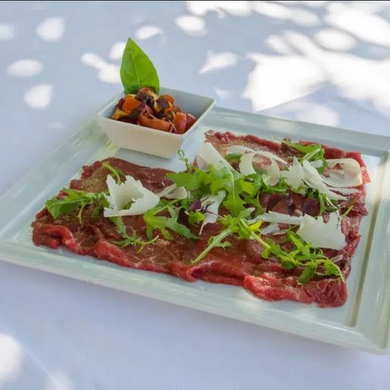 Ein Gericht aus Rindfleisch-Carpaccio mit Rucola, Parmesan und einer kleinen Beilage von Gemüse ist kunstvoll auf einem weißen Teller arrangiert, vor einem sanften, hellen Hintergrund.