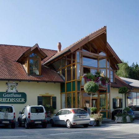 Das Gasthaus Friedisser hat ein rotes Ziegeldach und Glasfenster. Es hat zwei Topfpflanzen vor dem Gebäude. Vor dem Gebäude sind vier Autos geparkt.