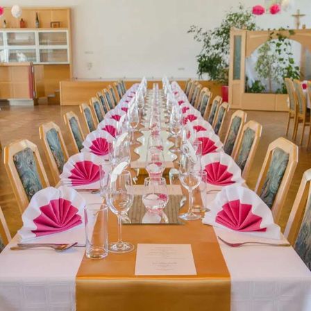 Ein langer Esstisch ist mit weißen Tischdecken, goldenen Tischläufern und rosa Servietten gedeckt. Weingläser und Besteck sind arrangiert. Stühle sind auf beiden Seiten platziert. Der Raum hat Holzböden und weiße Wände.