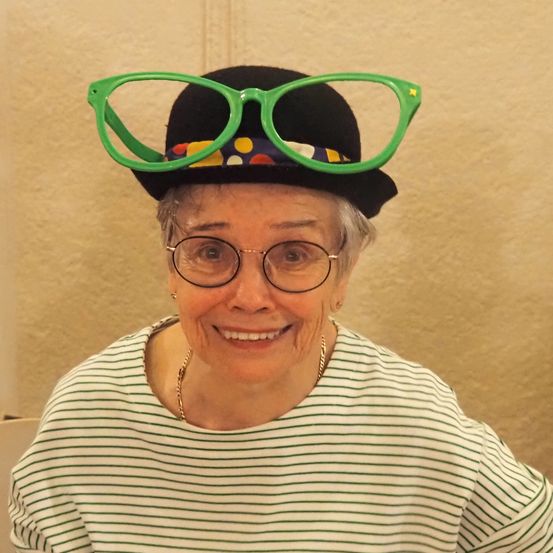Eine ältere Frau mit grauem Haar trägt einen schwarzen Hut mit grünen Brillengläsern obenauf und ein grün-weiß gestreiftes Hemd und lächelt in die Kamera.