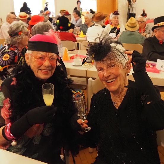 Zwei ältere Frauen, die Hüte und Handschuhe tragen, stoßen mit Champagnergläsern in einem überfüllten, festlichen Saal mit verschiedenen Dekorationen und anderen Gästen an.