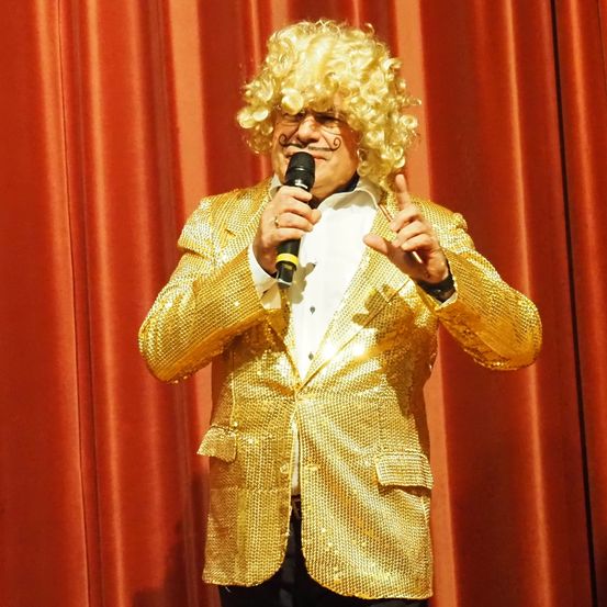Ein Mann mit lockigem blondem Haar und einem Schnurrbart trägt einen goldfarbenen Pailletten-Anzug und hält ein Mikrofon. Er steht auf einer Bühne mit roten Vorhängen dahinter.