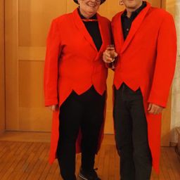 Zwei Menschen stehen nebeneinander und tragen rote Mäntel. Sie halten ein Glas Champagner und lächeln. Sie scheinen für ein Foto zu posieren.