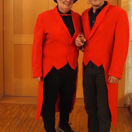 Zwei Menschen stehen nebeneinander und tragen rote Mäntel. Sie halten ein Glas Champagner und lächeln. Sie scheinen für ein Foto zu posieren.