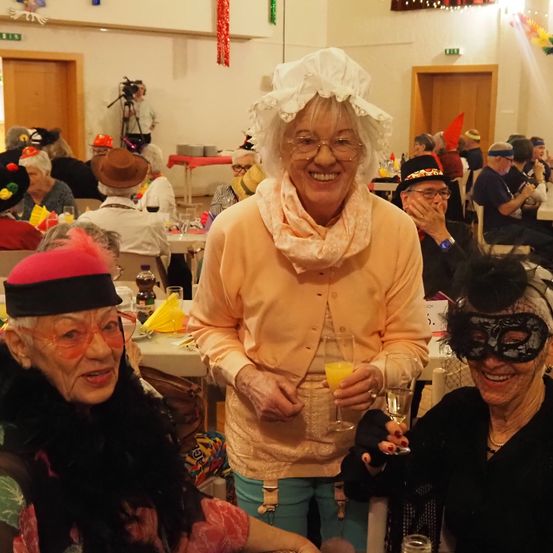 Mehrere ältere Frauen sind auf einer Party, wobei eine Frau in einem Kopftuch und einer Schürze ein Glas Orangensaft hält. Eine andere Frau trägt eine Maske und hält ein Glas Wein. Sie lächeln alle und posieren für ein Foto.