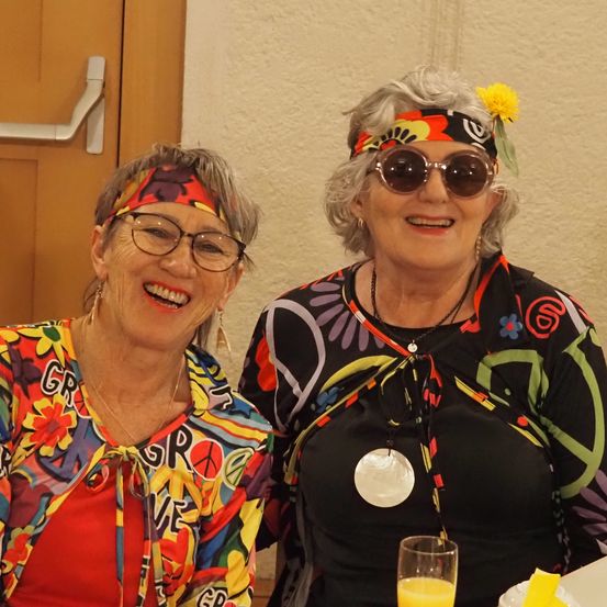 Zwei Frauen mit Sonnenbrillen und Kopfbändern mit Blumen sitzen eng beieinander und lächeln für ein Foto. Eine trägt eine bunte Jacke mit 'GR' und 'VE', die andere ein schwarzes Shirt mit einer Halskette. Dahinter steht eine Tür und ein Tisch mit einem Glas Orangensaft.