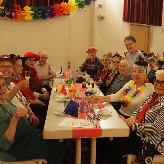 Eine Gruppe älterer Menschen in Hüten sitzt um einen Tisch herum, hält Gläser mit Wein und lächelt. An der Wand hinter ihnen hängen bunte Dekorationen.