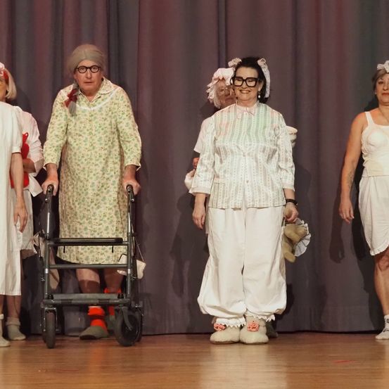 Fünf ältere Frauen in Vintage-Kleidung stehen auf der Bühne, eine benutzt einen Gehstock. Sie lächeln und stehen vor einem Vorhang.