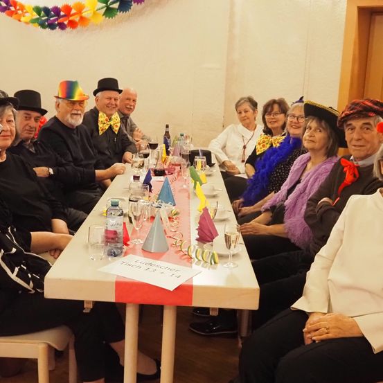 Eine Gruppe älterer Menschen sitzt um einen Tisch herum, trägt Hüte und bunte Schals, mit Weingläsern und Partydekoration.