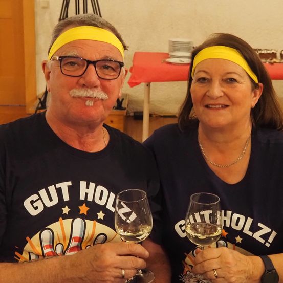 Ein Mann und eine Frau mit gelben Stirnbändern und passenden T-Shirts, die 'Gut Holz' sagen, halten Weingläser. Der Mann hat einen Schnurrbart und Brille, und die Frau lächelt. Hinter ihnen steht ein Tisch mit Tellern, und rechts ist ein Kuchen.