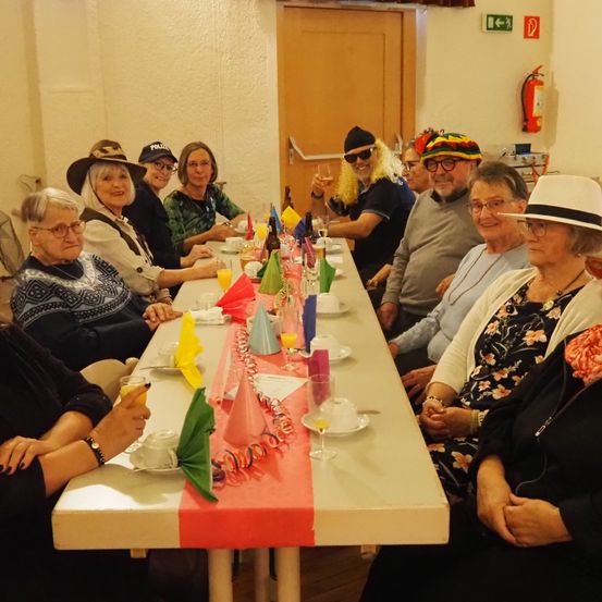 Eine Gruppe von Menschen, die verschiedene Hüte tragen, sitzt an einem langen Tisch und feiert. Sie haben Weingläser, Flaggen und bunte Servietten.