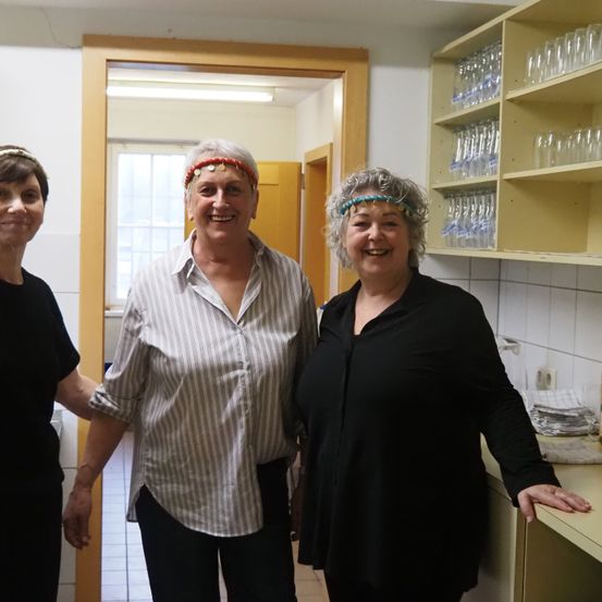 Drei Frauen stehen zusammen in einer Küche. Sie lächeln und tragen Stirnbänder. Hinter ihnen führt eine Tür in einen anderen Raum. Sichtbar sind Regale mit Gläsern und eine Theke mit einem Handtuch und Tellern.