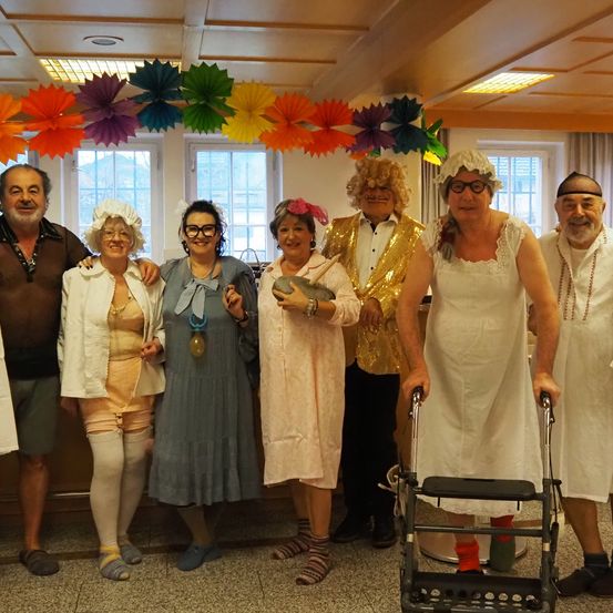 Eine Gruppe von Menschen in Kostümen posiert für ein Foto, mit bunten Papierdekorationen über ihnen. Eine Person benutzt einen Gehwagen und sie alle lächeln für die Kamera.