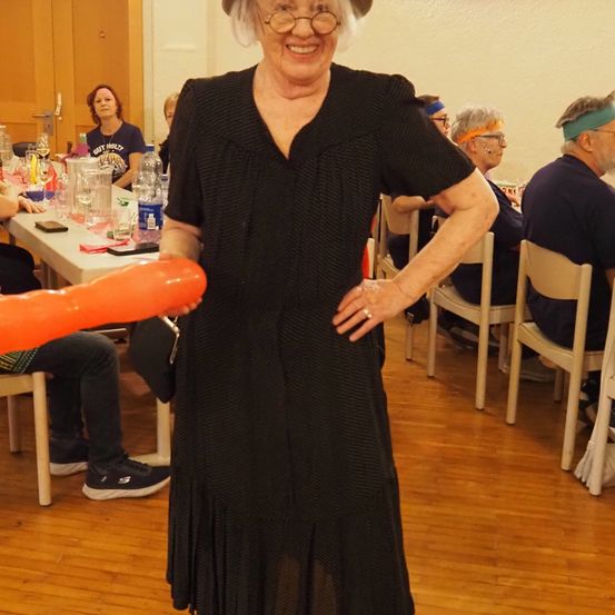 Eine ältere Frau in einem schwarzen Kleid steht in einem Raum mit anderen, die an einem Tisch sitzen, und hält ein großes orangefarbenes Objekt.