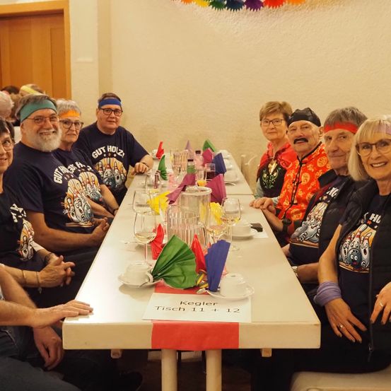 Eine Gruppe von Menschen sitzt mit passenden Stirnbändern und T-Shirts um einen Tisch. Der Tisch ist mit Gläsern, Tellern und Tassen gedeckt. Ein Schild steht 'Kegler Tisch 11 + 12'.