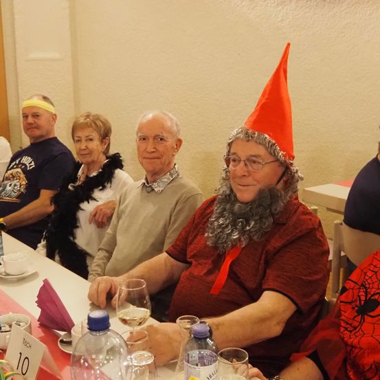 Eine Gruppe von Menschen sitzt an einem Tisch, darunter ein älterer Mann in einem roten Kostüm und Hut, mit Brille und Bart. Ein anderer Mann mit gelbem Stirnband und eine Frau in Schwarz sind ebenfalls anwesend.