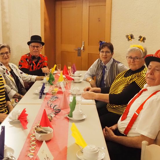 Eine Gruppe älterer Erwachsener in bunten Kostümen sitzt um einen langen Tisch herum, wahrscheinlich auf einer Party. Der Tisch ist mit Tellern, Tassen und Servietten gedeckt. Es gibt eine rote Tischdecke auf dem Tisch und eine Person trägt einen roten Hut.