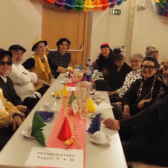 Eine Gruppe von Menschen sitzt bei einer Party um einen Tisch, alle in bunten Kostümen gekleidet. Sie lächeln und schauen in die Kamera. Der Tisch ist mit Weingläsern, Tassen, Tellern und Servietten gedeckt. Ein Whiteboard mit dem Text 'Mitwirkende Tisch 7 + 8' steht auf dem Tisch. Der Raum ist mit bunten Papierdekorationen an der Decke geschmückt.