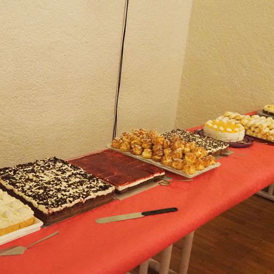Ein langer Tisch, der mit einem roten Tuch bedeckt ist und verschiedene Desserts zeigt, darunter Kuchen, Gebäck und schokoladenüberzogene Artikel. Die Desserts sind auf weißen Tellern angeordnet.