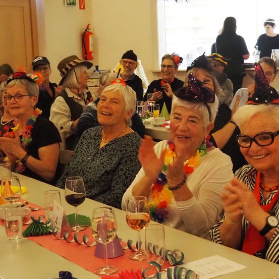 Eine Gruppe älterer Frauen mit festlichen Hüten und Brillen sitzt um einen Tisch mit Weingläsern, lächelt und klatscht. Einige tragen Halsketten. Im Hintergrund stehen Personen und ein Feuerlöscher ist an der Wand montiert.