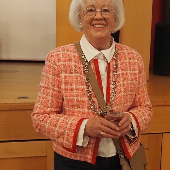 Eine ältere Frau in einem roten und weißen karierten Blazer, einem weißen Hemd und einer Kette mit einer roten Schleife lächelt und hält ein Getränk in der Hand.