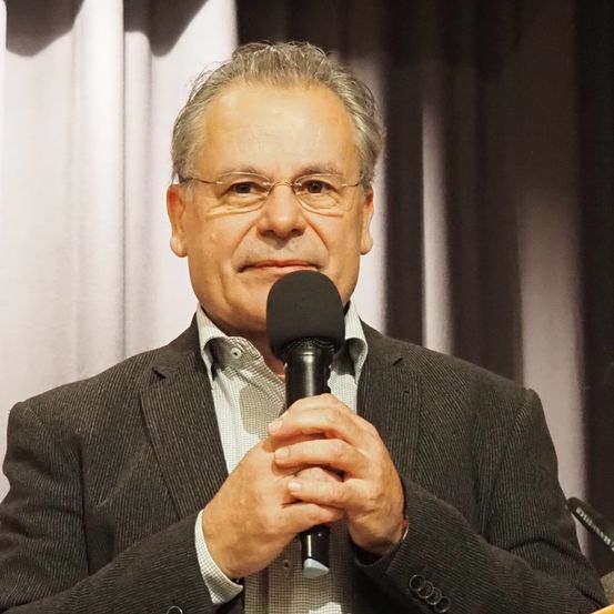 Ein Mann mit grauen Haaren, der eine Brille und einen schwarzen Anzug trägt, spricht in ein Mikrofon. Er steht vor einem weißen Vorhang.