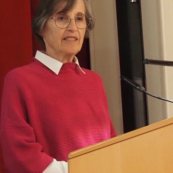 Eine ältere Frau mit grauem Haar, die eine Brille und einen roten Pullover trägt, spricht an einem Rednerpult. Ein Mikrofon steht vor ihr.
