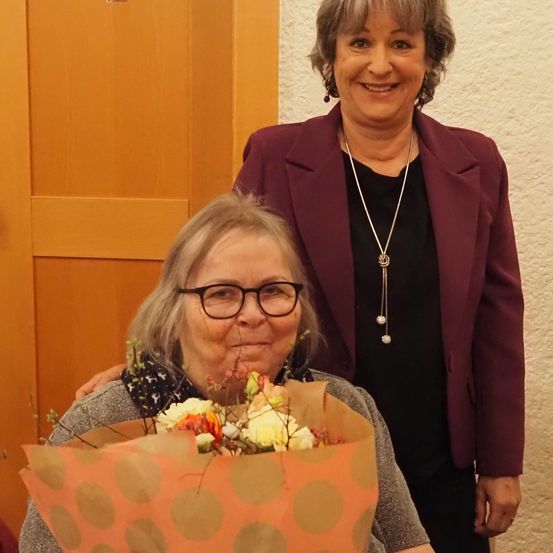 Zwei Frauen stehen zusammen; eine hält einen Blumenstrauß, die andere hat eine Halskette und trägt eine weinrote Blazerjacke. Hinter ihnen ist eine Holztür.