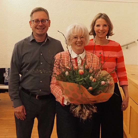 Drei Menschen stehen eng beieinander und lächeln. Die Frau in der Mitte hält einen Blumenstrauß. Der Mann auf der linken Seite trägt eine Brille und einen Gürtel, und die Frau auf der rechten Seite trägt ein gestreiftes Hemd. Sie stehen in einem Raum mit einer weißen Wand.