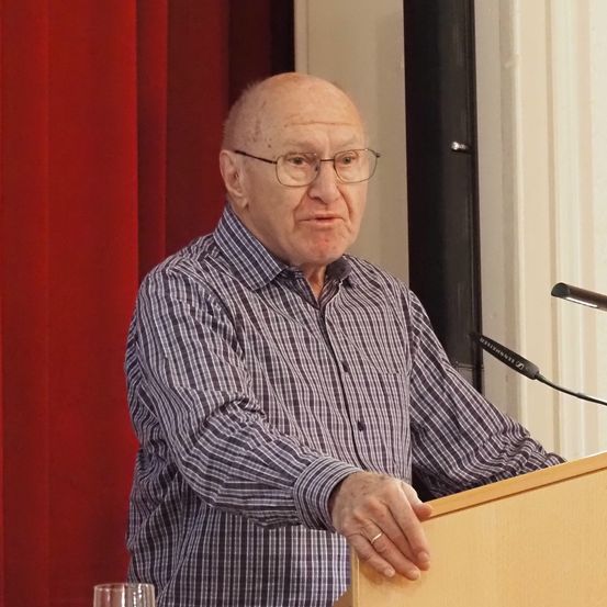 Ein älterer Mann mit Brille und kariertem Hemd spricht an einem Podium mit einem Mikrofon vor ihm.
