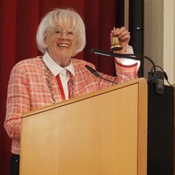 Eine ältere Frau, lächelnd, steht an einem Rednerpult und hält eine Glocke in der Hand, trägt eine Brille und ein rot-weiß kariertes Sweatshirt.