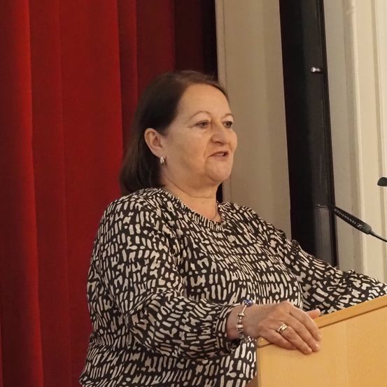 Eine Frau steht an einem Rednerpult, trägt ein gemustertes Oberteil, eine Halskette und Ringe. Sie lächelt das Publikum an, mit einem roten Vorhang und einem Mikrofon im Hintergrund.