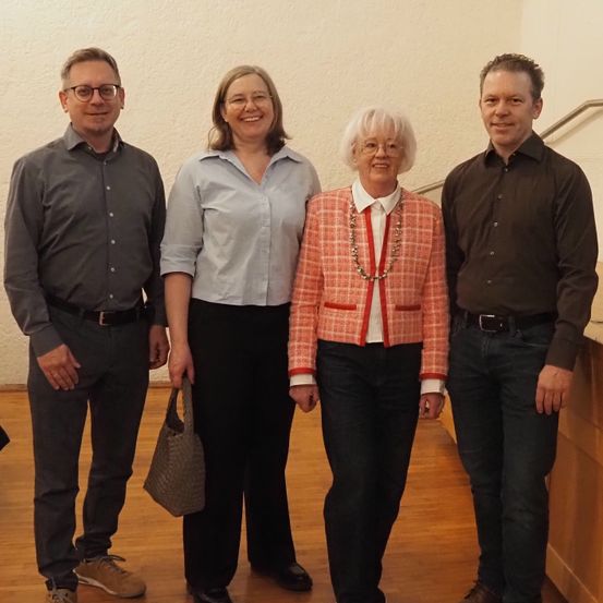 Vier Personen stehen in einem Raum mit Holzboden. Zwei Männer und zwei Frauen. Sie lächeln für ein Foto. Die Frau in der Mitte trägt eine Halskette. Die Männer tragen Gürtel und eine Brille.