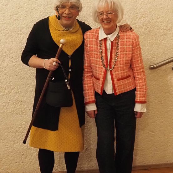 Zwei ältere Frauen in Kostümen, eine mit einem Stock, posieren vor einer Wand.