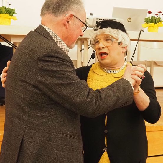 Ein älterer Mann im Anzug tanzt mit einer älteren Frau in einem gelben Kleid und schwarzem Hut, mit einem Laptop und Blumen im Hintergrund.