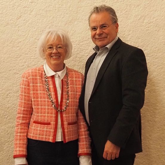 Eine ältere Frau in einer roten Jacke mit einer Halskette steht neben einem Mann in einem schwarzen Anzug, beide lächeln und posieren für ein Foto vor einer weißen Wand.